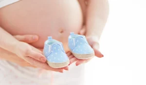 Eine schwangere Frau hält ein Paar kleine, blaue Babyschuhe mit weißen Punkten in ihren Händen vor ihrem nackten Babybauch. Ihre Hände stützen den Bauch, während die Schuhe sanft präsentiert werden. Das Bild vermittelt Vorfreude auf die bevorstehende Geburt.