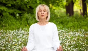 Eine Frau mittleren Alters sitzt in einem weißen Outfit auf einer Wiese, umgeben von kleinen weißen Blumen. Sie hat die Augen geschlossen und hält ihre Hände in einer Meditationshaltung. Der Hintergrund zeigt üppiges Grün, das eine ruhige und friedliche Atmosphäre schafft. Die Frau scheint in tiefer Entspannung und Achtsamkeit vertieft zu sein.